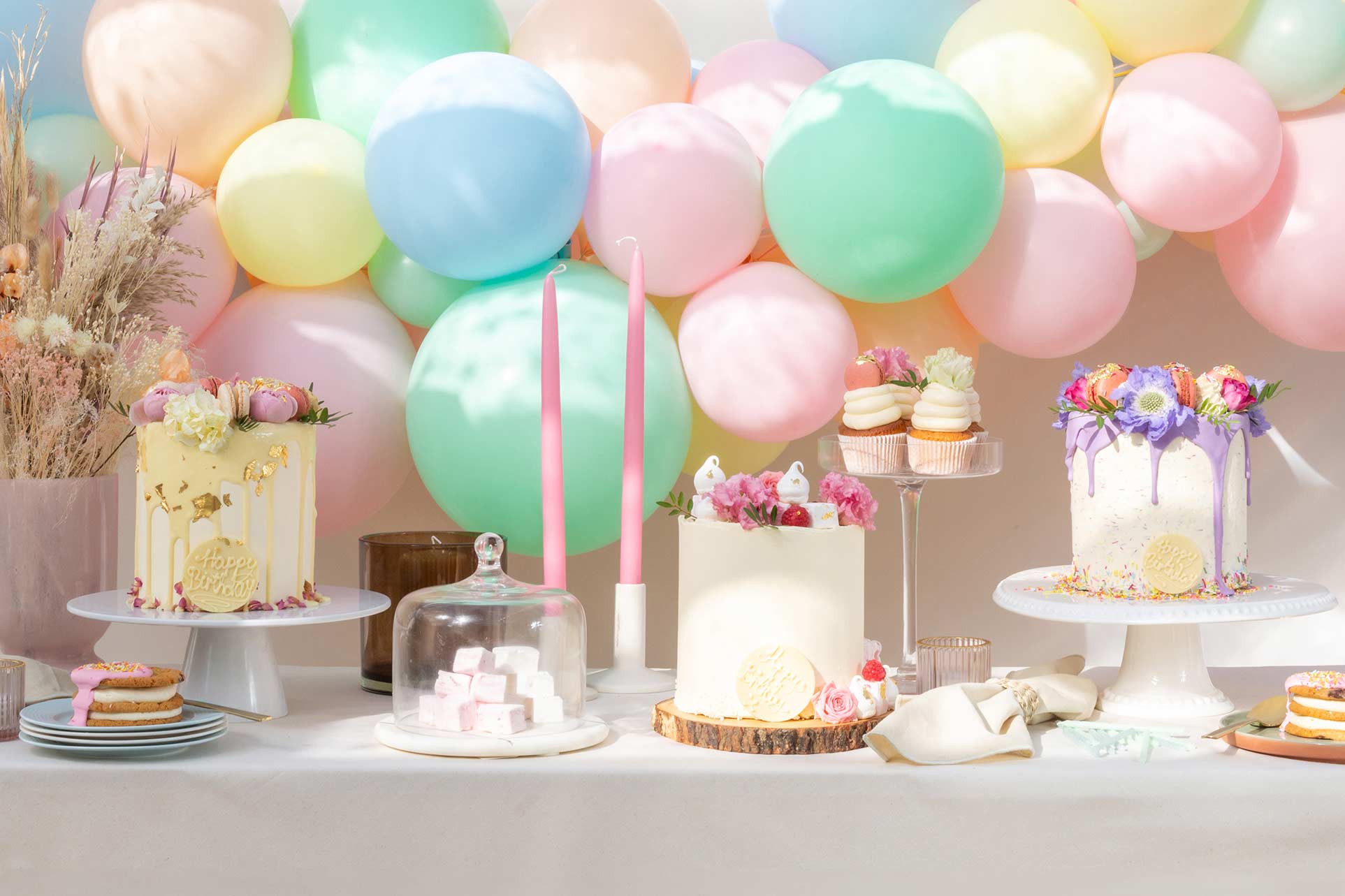 Celebration table with three cakes, lots of sweet treats such as cup cakes surrounded by balloons